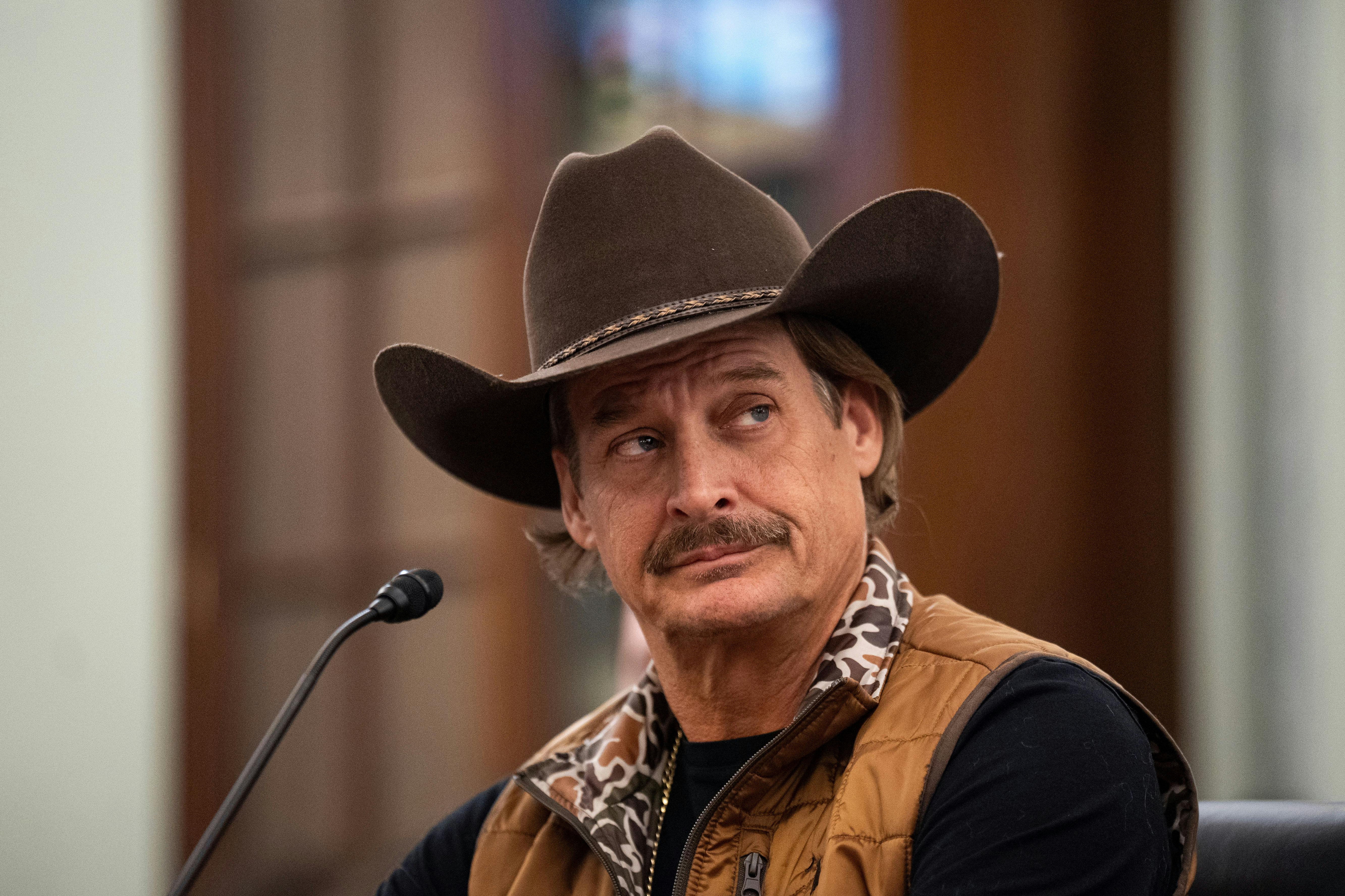 Kid Rock looks to the side while sitting at a microphone in a congressional hearing