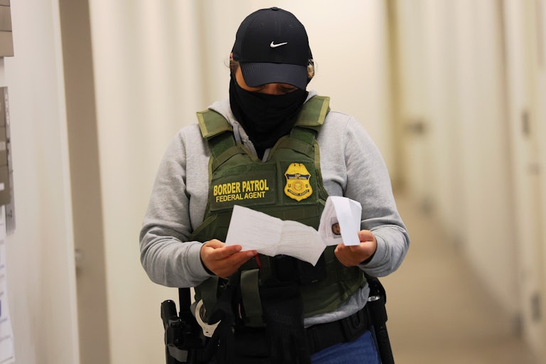 A masked ICE agent wearing a cap and a Border Patrol vest reads a piece of paper in his hands.