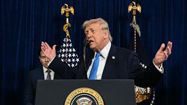 Donald Trump holds both hands out in a shrug-like gesture while standing at a podium, with Marco Rubio in the background.