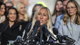 Marjorie Taylor Greene speaks at a lectern with lots of mics, as survivors of Jeffrey Epstein's abuse stand in the background.