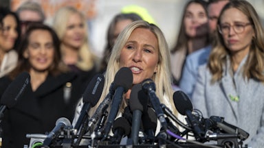 Marjorie Taylor Greene speaks at a lectern with lots of mics, as survivors of Jeffrey Epstein's abuse stand in the background.