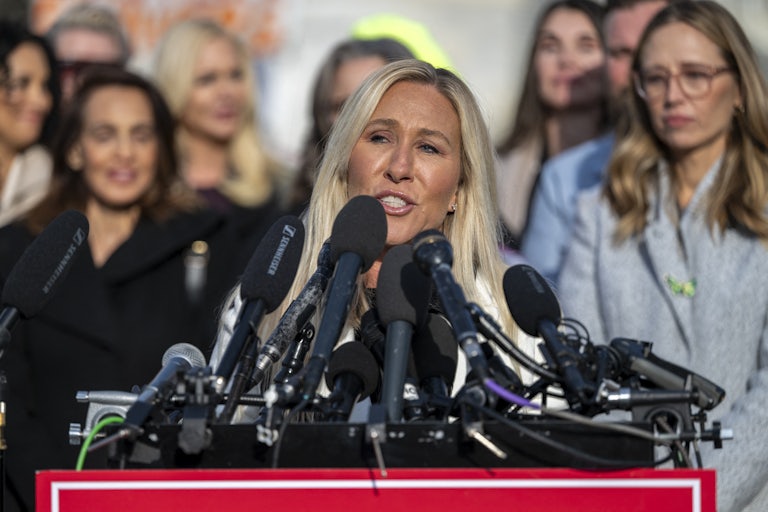 Marjorie Taylor Greene speaks at a lectern with lots of mics, as survivors of Jeffrey Epstein's abuse stand in the background.