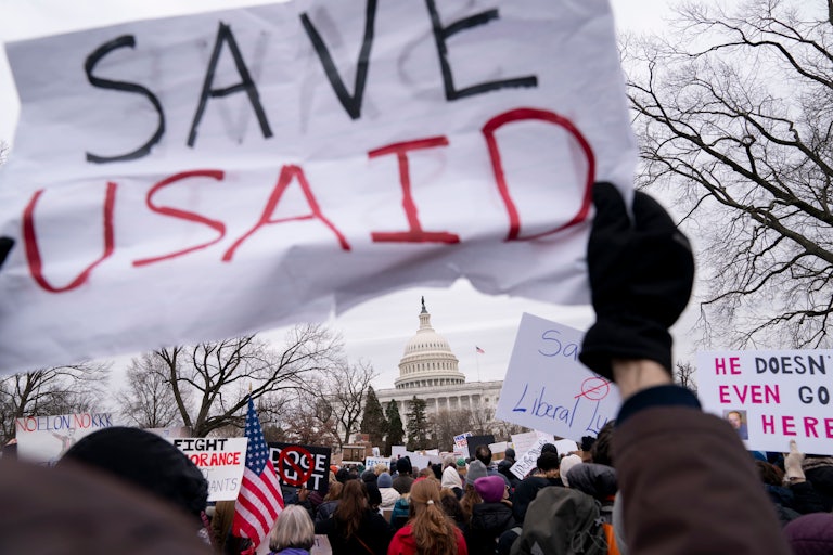 A crowd of protesters outside the Capitol hold up signs. One in the foreground reads "SAVE USAID."