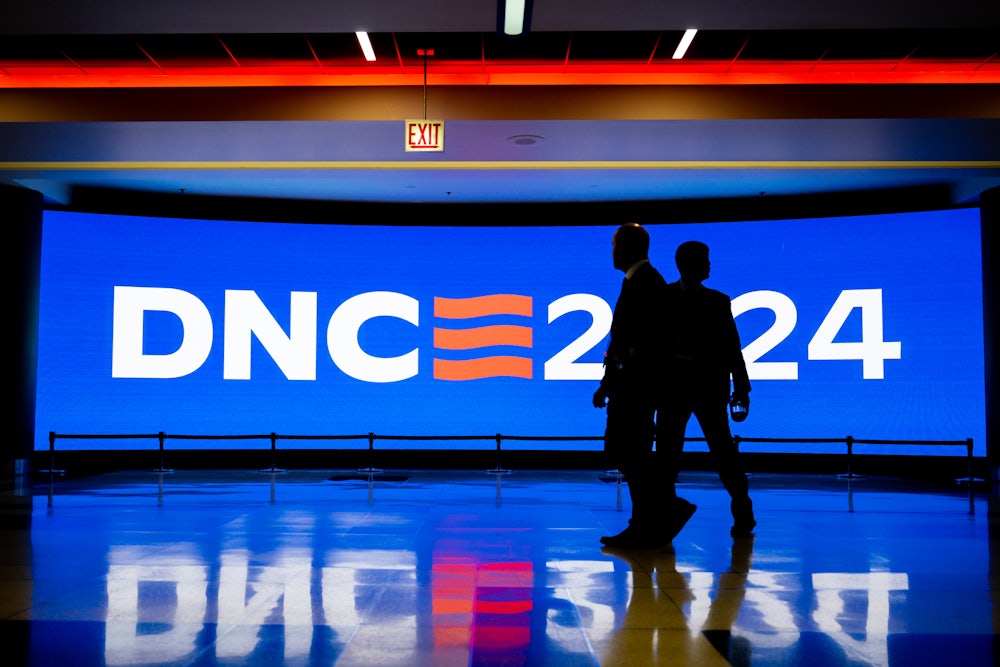 Two security officials are seen in silhouette walking in front of a bright blue screen that reads "DNC 2024."