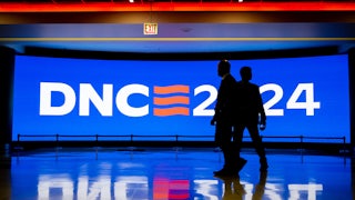 Two security officials are seen in silhouette walking in front of a bright blue screen that reads "DNC 2024."