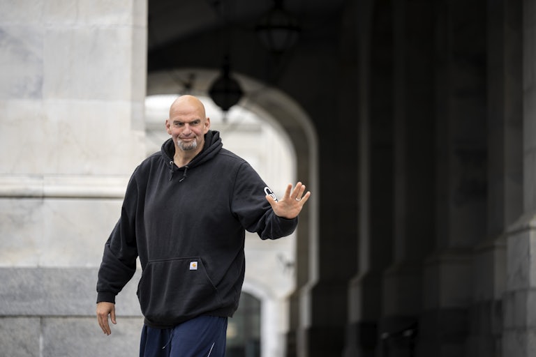 John Fetterman wearing a hoodie smiles and waves at reporters as he arrives at the Capitol