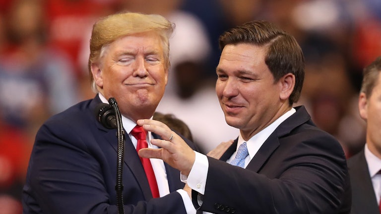 Donald Trump and Ron DeSantis exchange greetings behind a lectern at a rally.