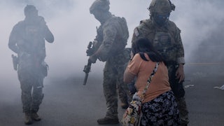 Three masked federal agents with guns walk in tear gas as a woman tries to cover her face.