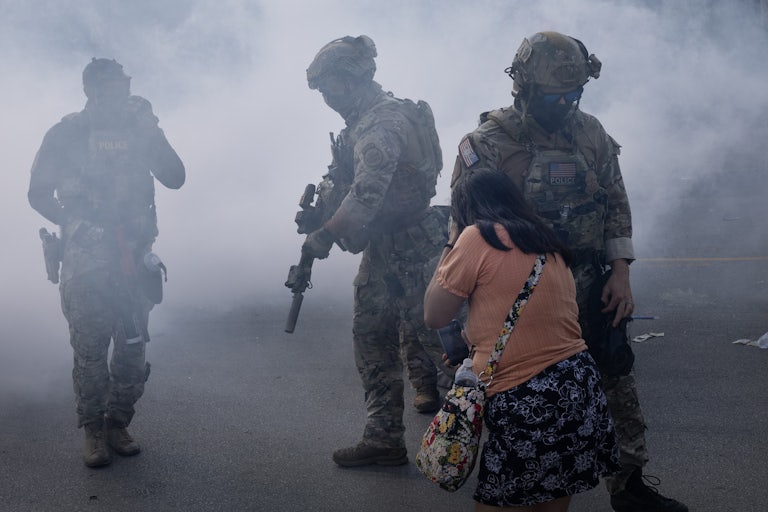 Three masked federal agents with guns walk in tear gas as a woman tries to cover her face.