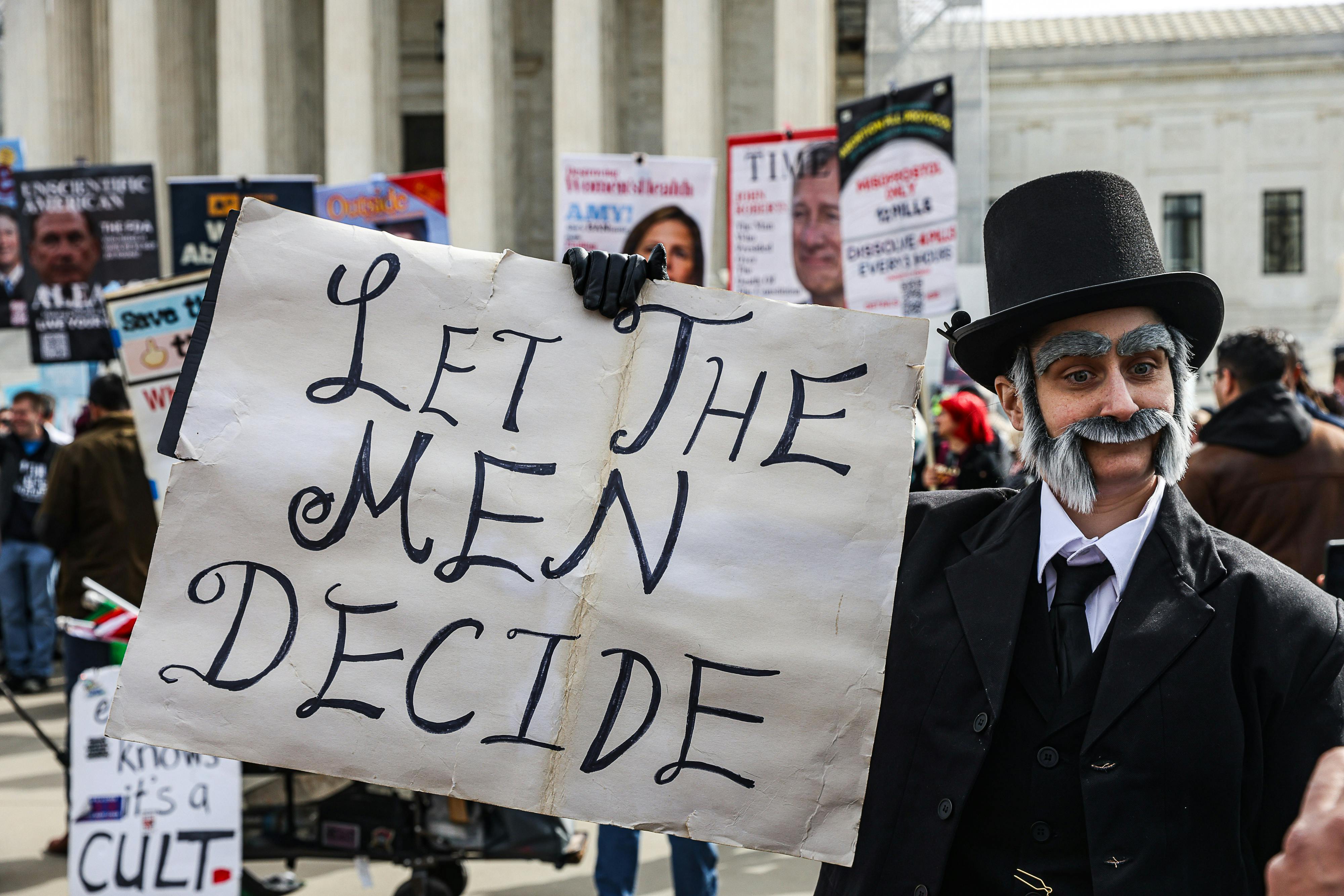 A demonstrator in historical costume holds a sign reading "let the men decide"