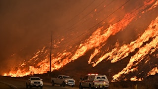 Emergency vehicles stand on the road in front of a hill on fire.