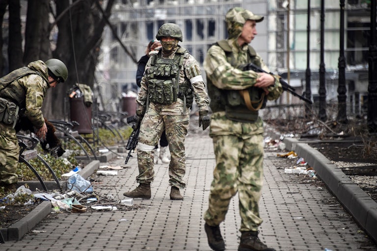 Three soldiers carry guns in a street littered with detritus.