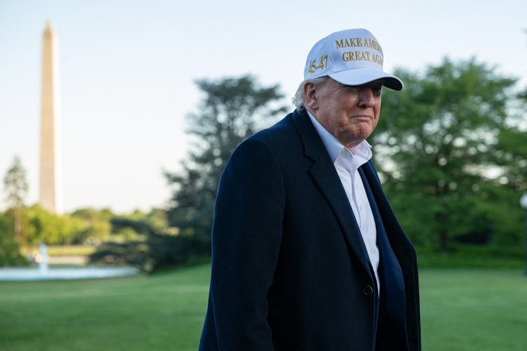 Donald Trump wears a white Make America Great Again hat while standing outside the White House. The Washington Monument is visible behind him