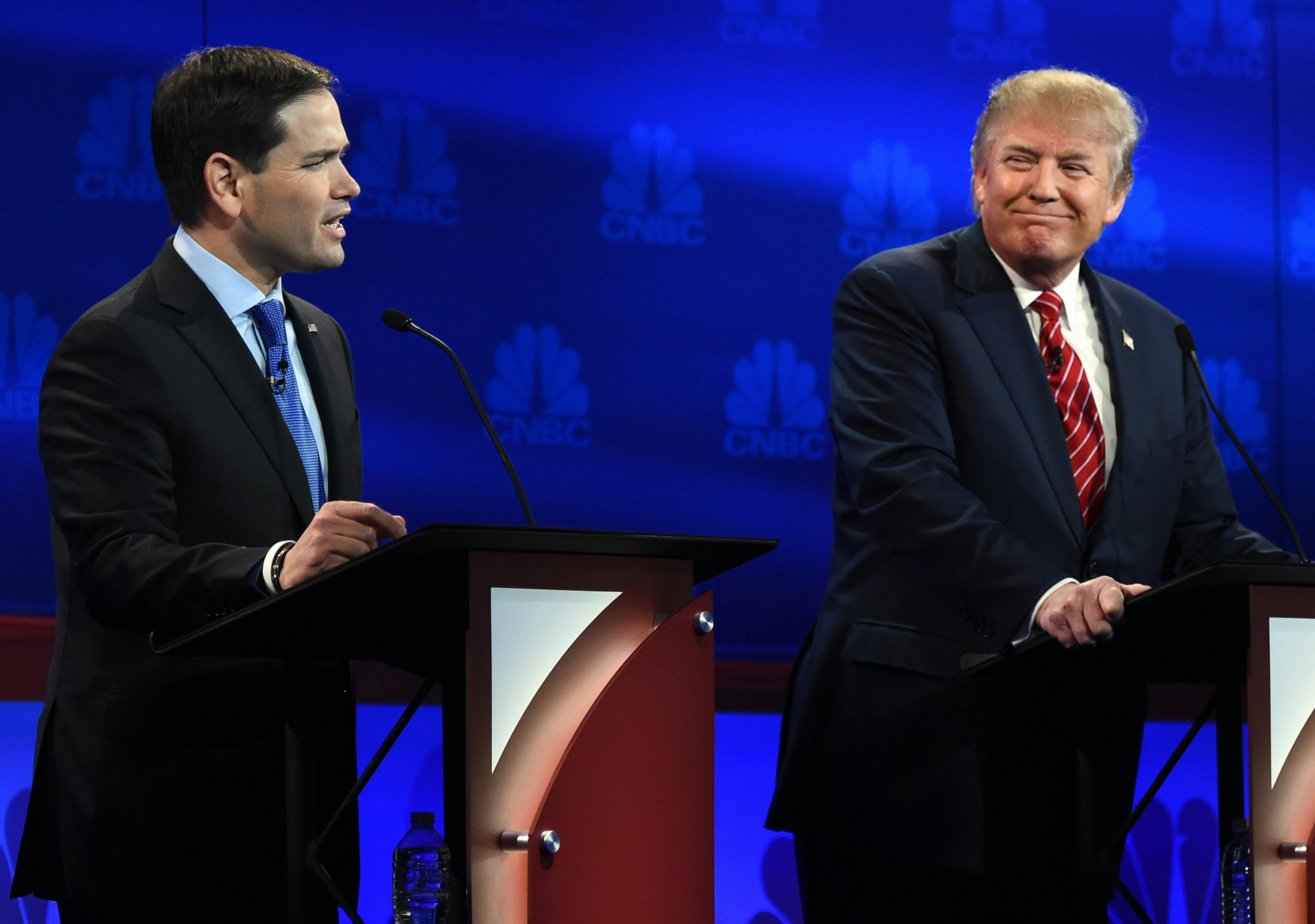Donald Trump smiles as he stands behind a lectern and looks over at Marco Rubio, who is talking. 
