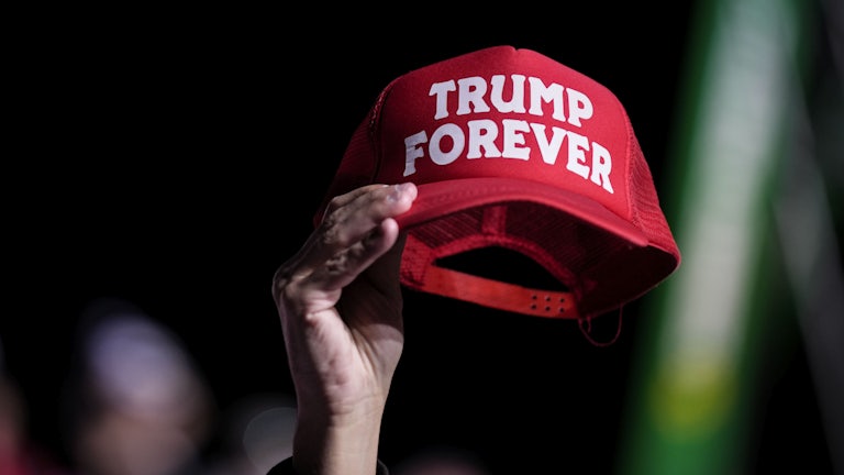 A hand holds a red hat reading "TRUMP FOREVER."