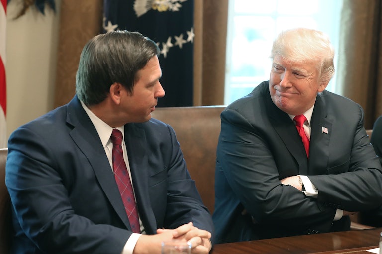 Ron DeSantis sits at a table next to Donald Trump who smiles smugly with his arms folded