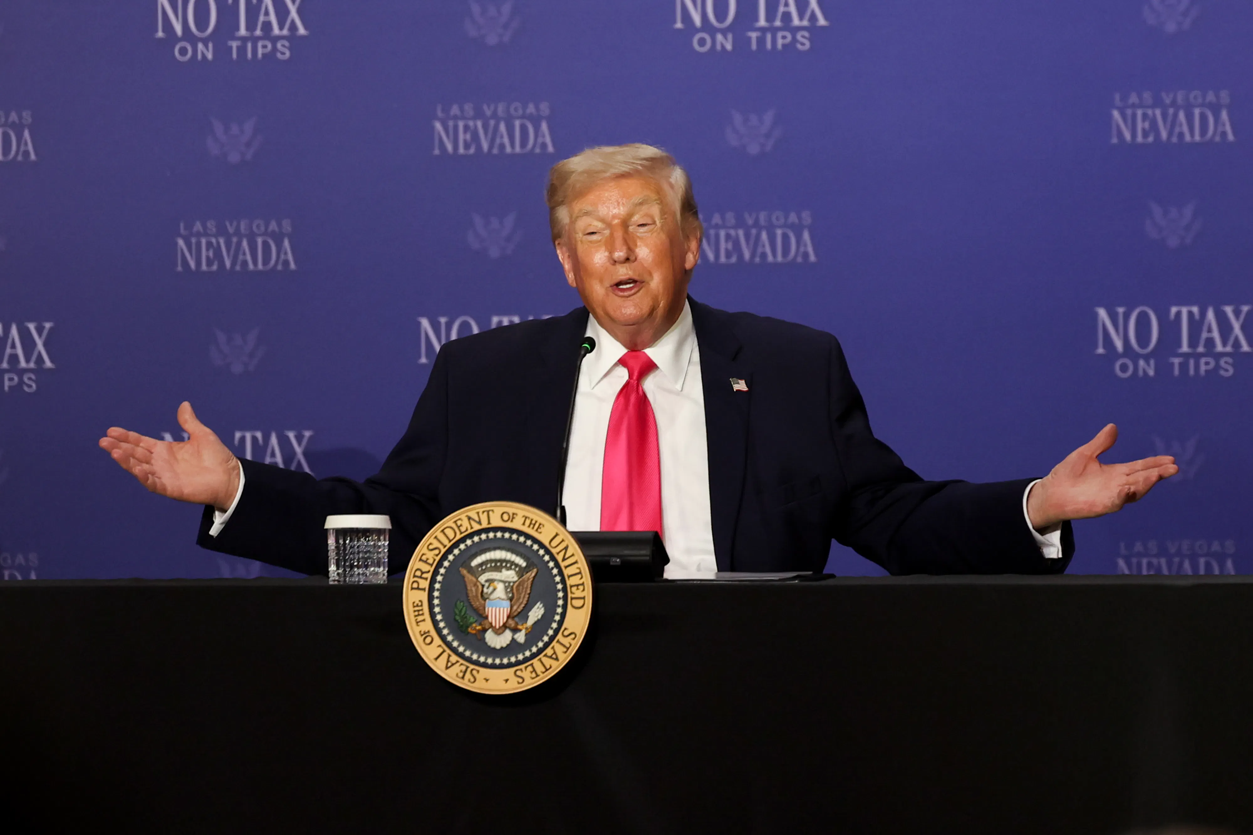 Donald Trump holds his arms out to the side while sitting at a table