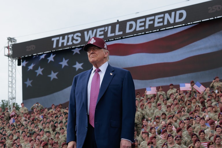 Donald Trump smiles while standing in front of troops. A sign at the top reads "This We'll Defend."