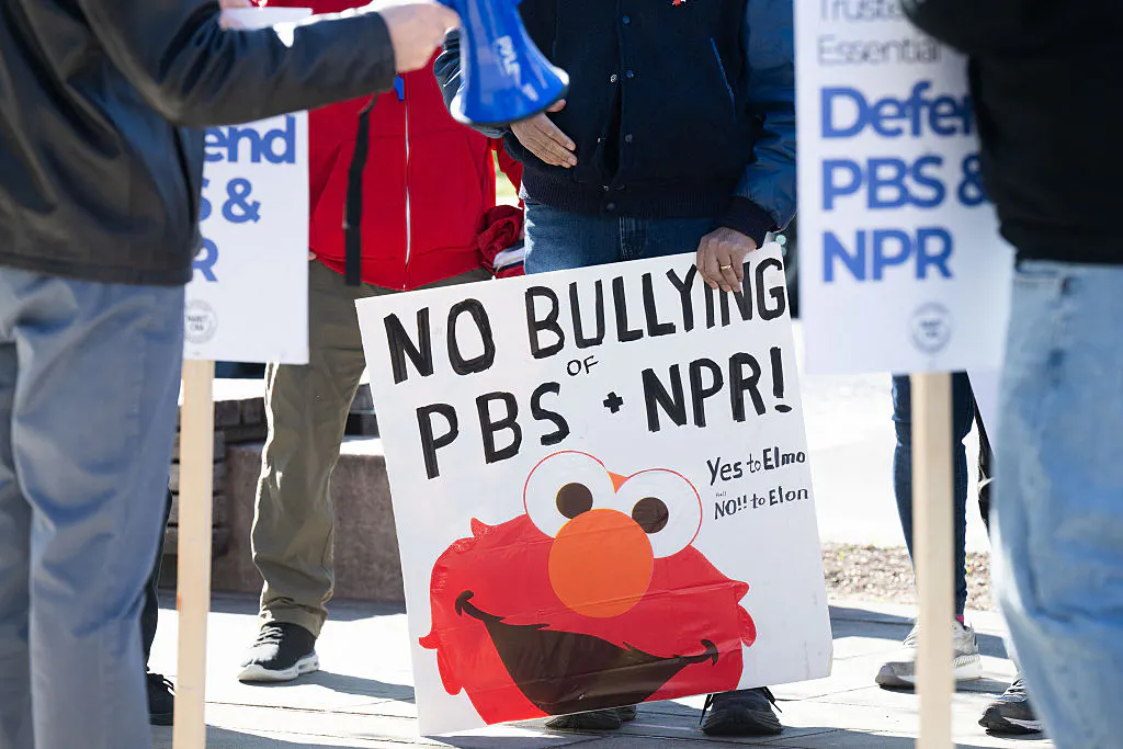Someone holds a sign that says "No Bullying of NPR + PBS" featuring an image of Elmo.