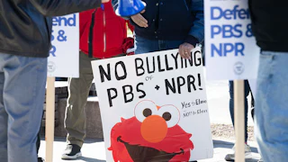 Someone holds a sign that says "No Bullying of NPR + PBS" featuring an image of Elmo.