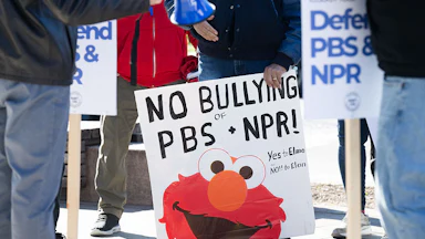 Someone holds a sign that says "No Bullying of NPR + PBS" featuring an image of Elmo.