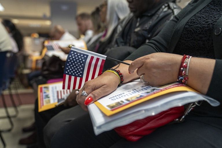 A brown woman seated holds a U.S. flag in her left hand and a stack of papers in her lap. The paper on top reads "Congratulations on Becoming a U.S. Citizen."