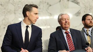 Josh Hawley looks to his left at Lindsey Graham, who is grimacing. Both are wearing blue suits and standing behind black leather chairs, and a Senate staffer is visible behind Graham to his left.