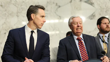 Josh Hawley looks to his left at Lindsey Graham, who is grimacing. Both are wearing blue suits and standing behind black leather chairs, and a Senate staffer is visible behind Graham to his left.