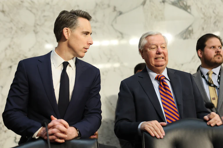Josh Hawley looks to his left at Lindsey Graham, who is grimacing. Both are wearing blue suits and standing behind black leather chairs, and a Senate staffer is visible behind Graham to his left.