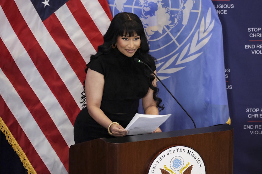 Nicki Minaj holds two pieces of paper while standing at a U.N. podium. The U.S. and U.N. flags are behind her.
