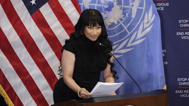 Nicki Minaj holds two pieces of paper while standing at a U.N. podium. The U.S. and U.N. flags are behind her.