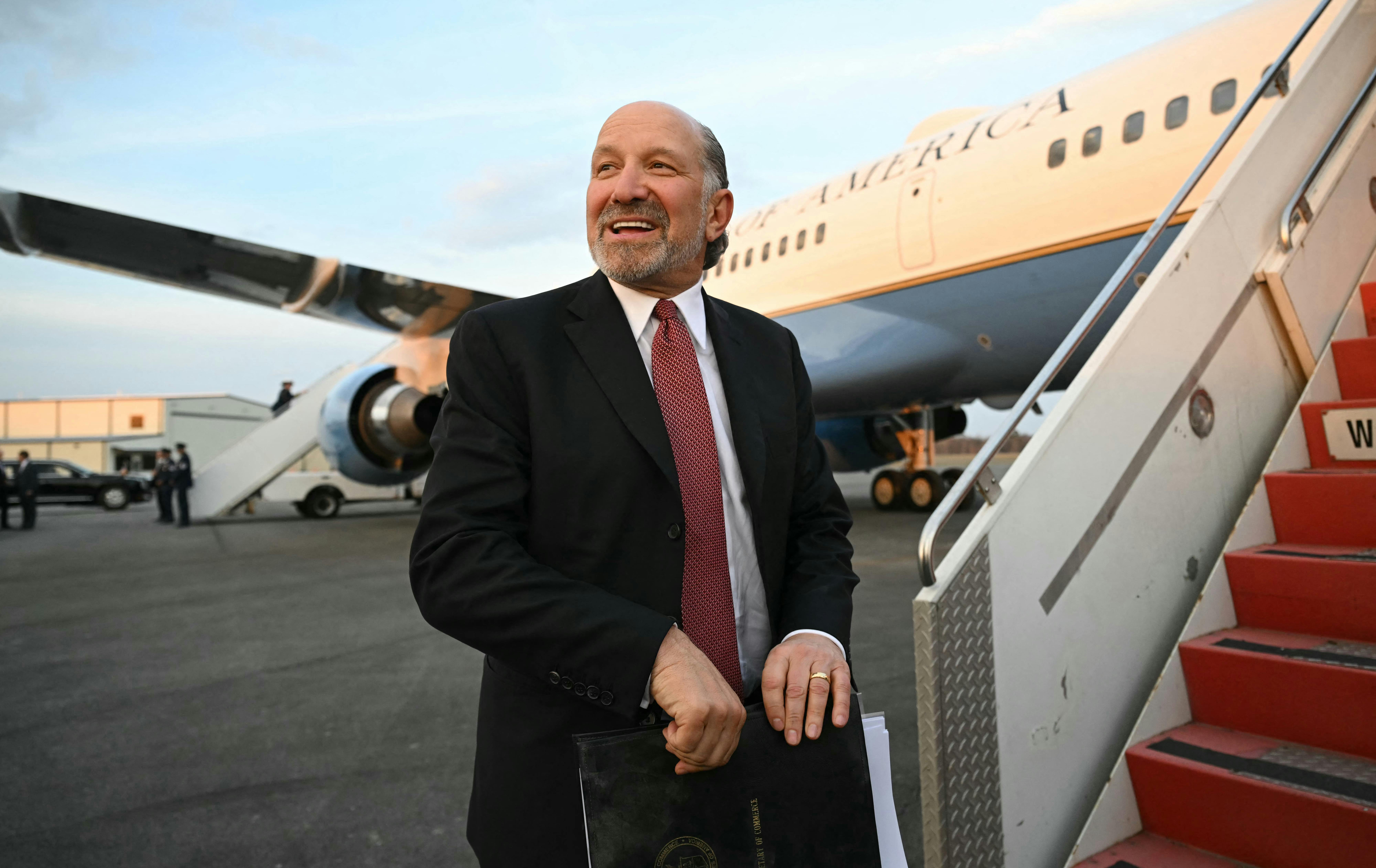 Commerce Secretary Howard Lutnick holds a folder and smiles as he boards Air Force One.