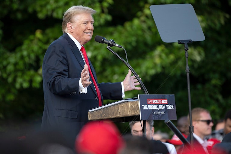 Donald Trump stands at a lectern on stage, splays his hands out and smiles. A few heads in a crowd (out of focus) are below him.