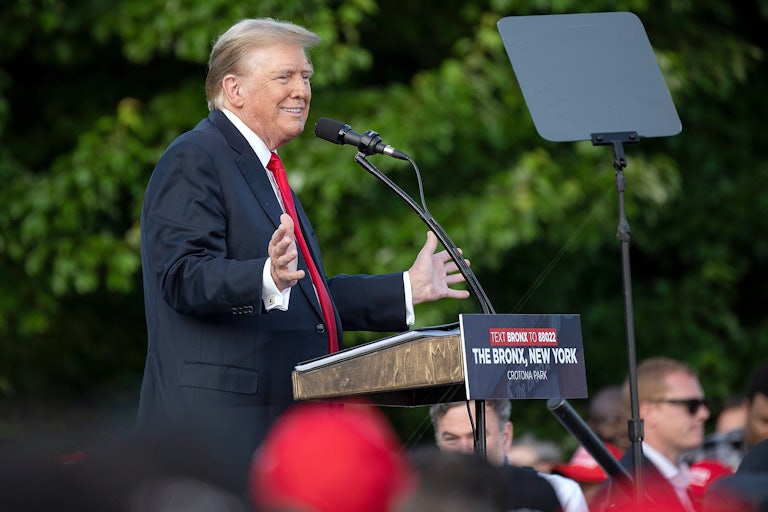 Donald Trump stands at a lectern on stage, splays his hands out and smiles. A few heads in a crowd (out of focus) are below him.