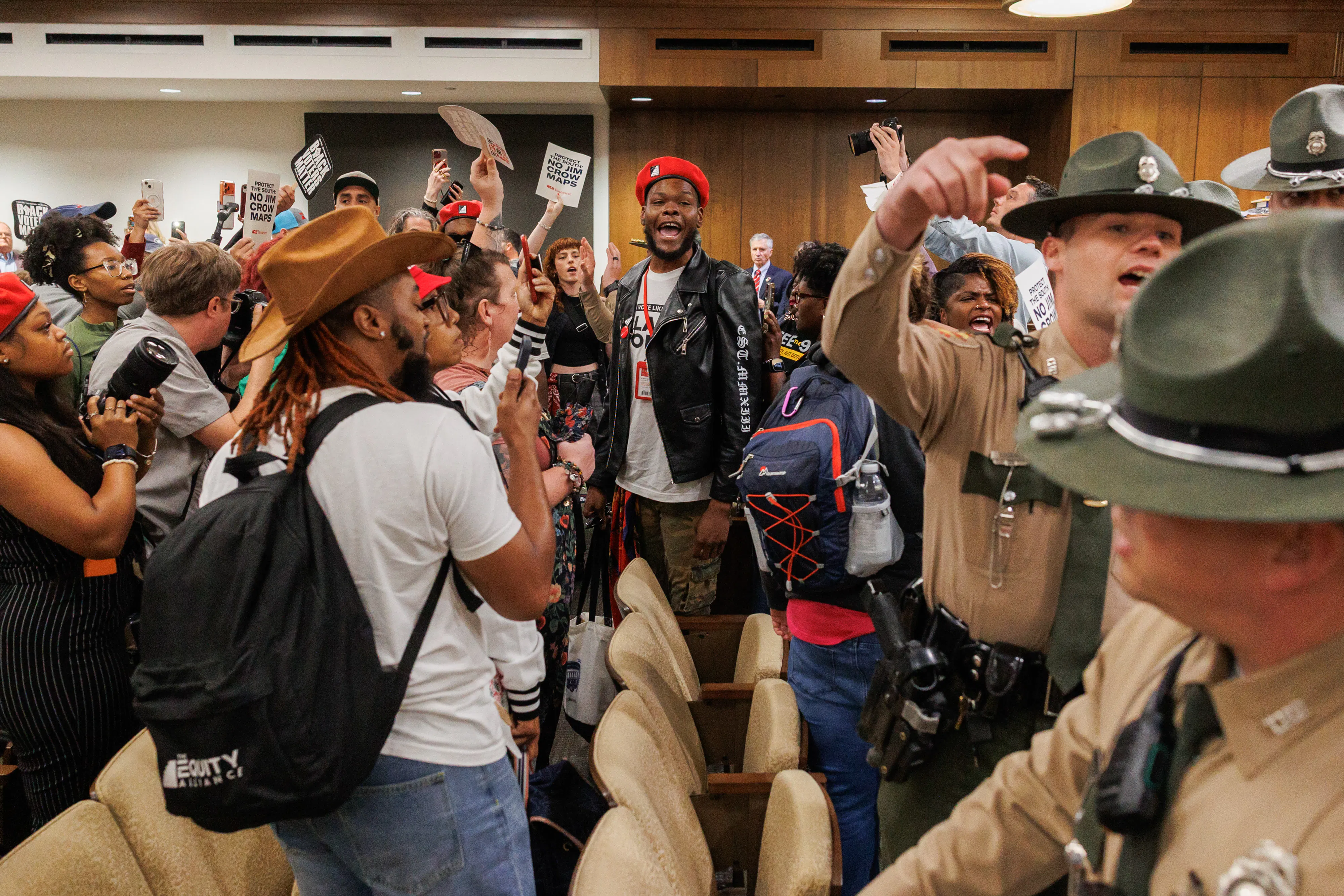 Attendees protest, yell, and hold signs opposed to gerrymandering as police stand nearby.