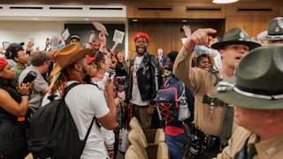 Attendees protest, yell, and hold signs opposed to gerrymandering as police stand nearby.