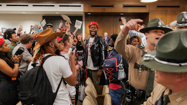 Attendees protest, yell, and hold signs opposed to gerrymandering as police stand nearby.