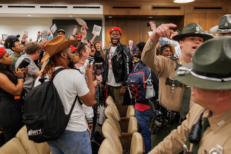Attendees protest, yell, and hold signs opposed to gerrymandering as police stand nearby.