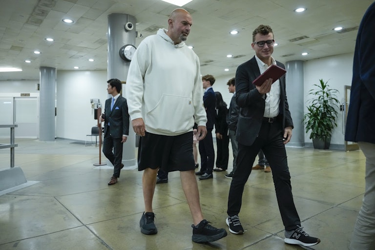 Senator John Fetterman wears shorts and talks to an aide who is wearing a suit and tie. They are walking through the Senate subway.