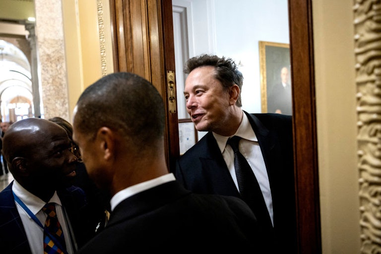 Elon Musk leans out of a doorway, smiling.