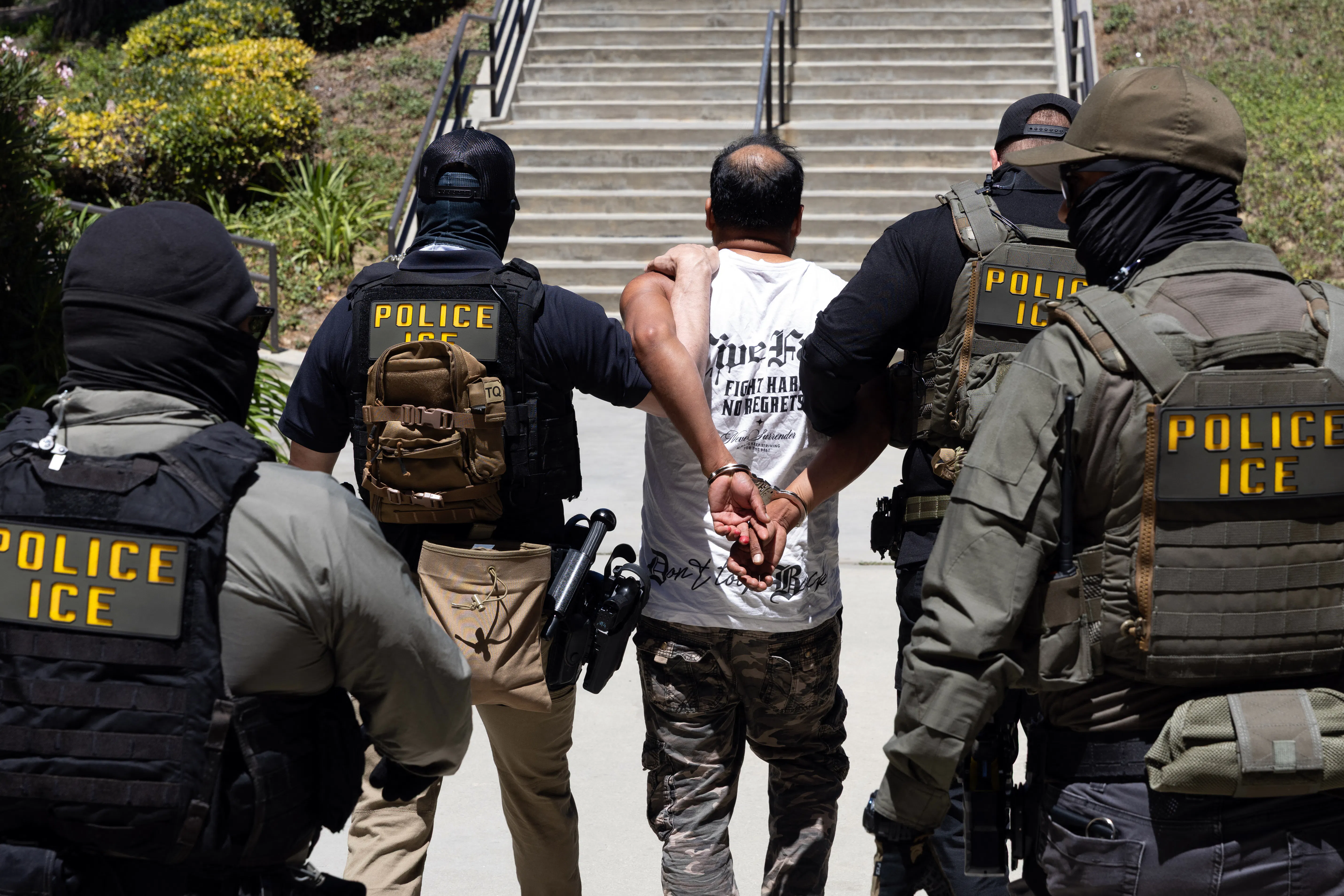Four masked ICE agents guide a handcuffed man