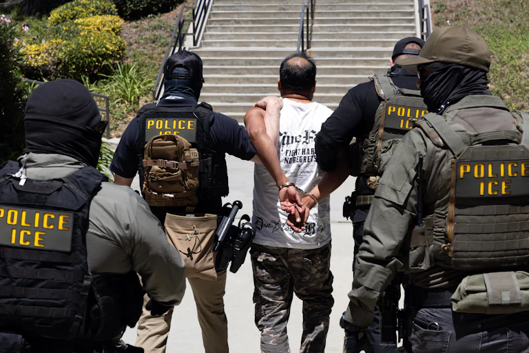 Four masked ICE agents guide a handcuffed man