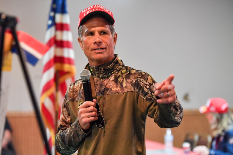 Dan Meuser wears camo while standing in front of an American flag holding a microphone and gesticulating.