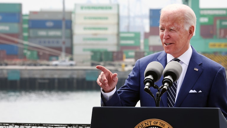 President Biden speaks at a podium, with a harbor and shipping containers visible behind him.