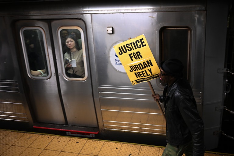 A woman riding the subway looks at a protestor carrying a “Justice for Jordan Neely” poster walking on the platform.