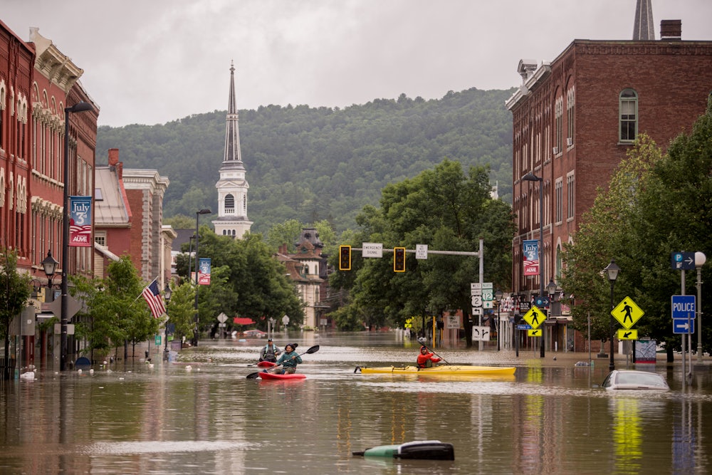 People kayak down a flooded city street; wooded mountains rise up in the background.