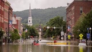 People kayak down a flooded city street; wooded mountains rise up in the background.