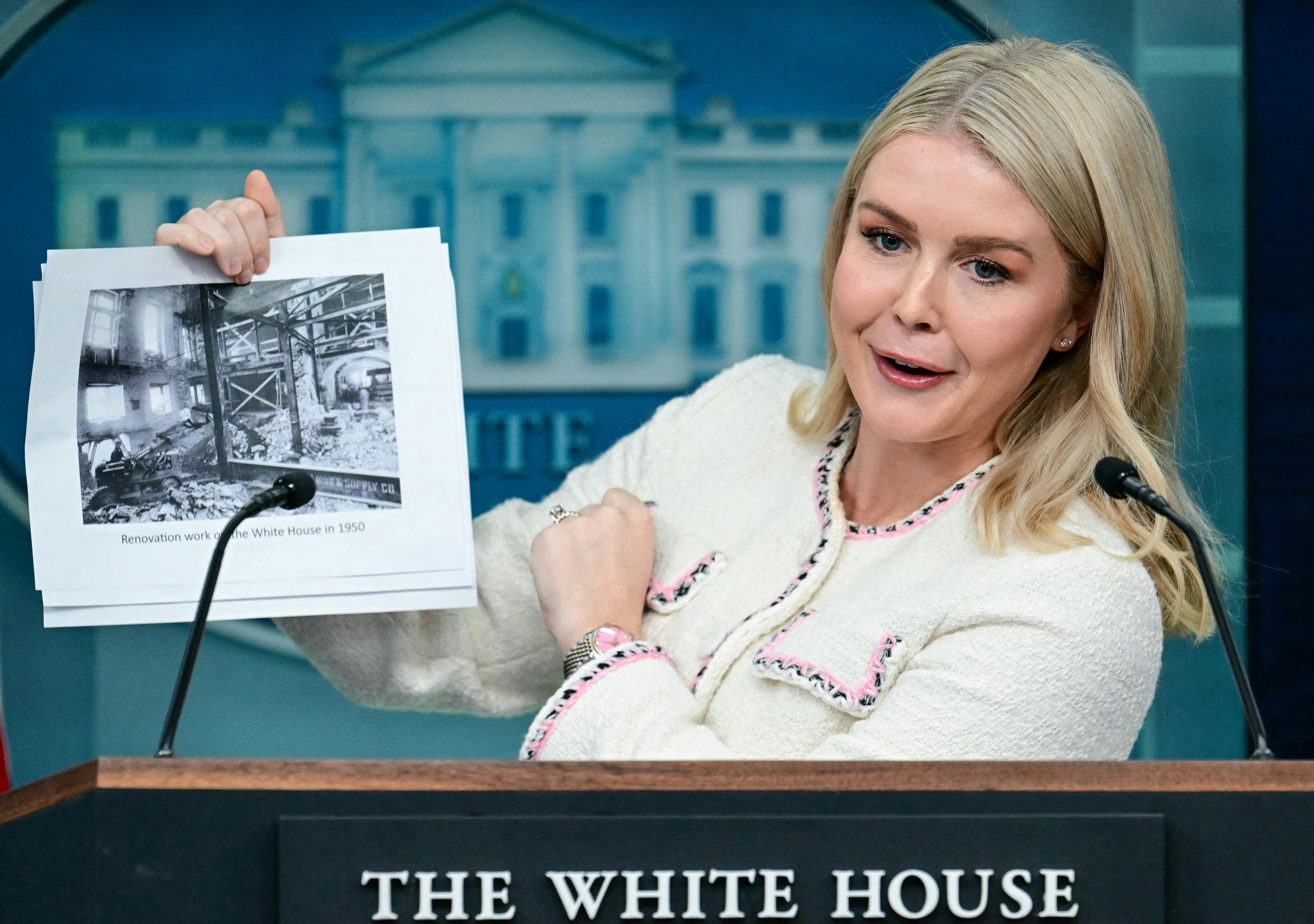 Karoline Leavitt speaks at the podium in the White House briefing room while holding up a photo of previous White House renovations