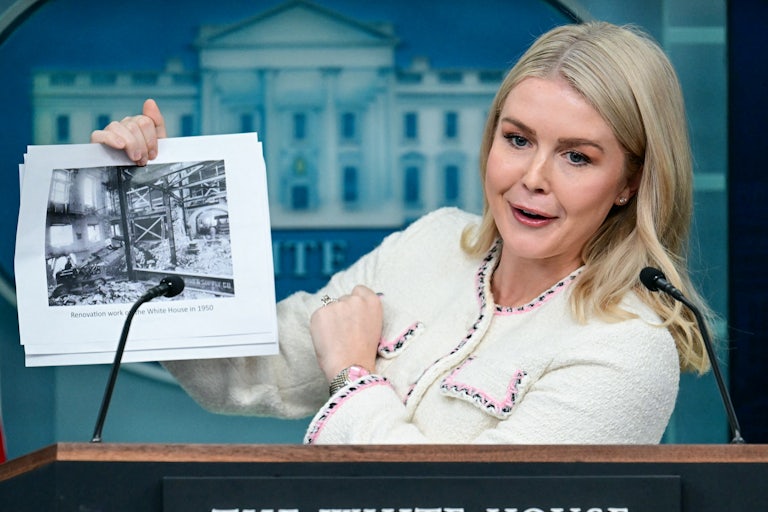 Karoline Leavitt speaks at the podium in the White House briefing room while holding up a photo of previous White House renovations