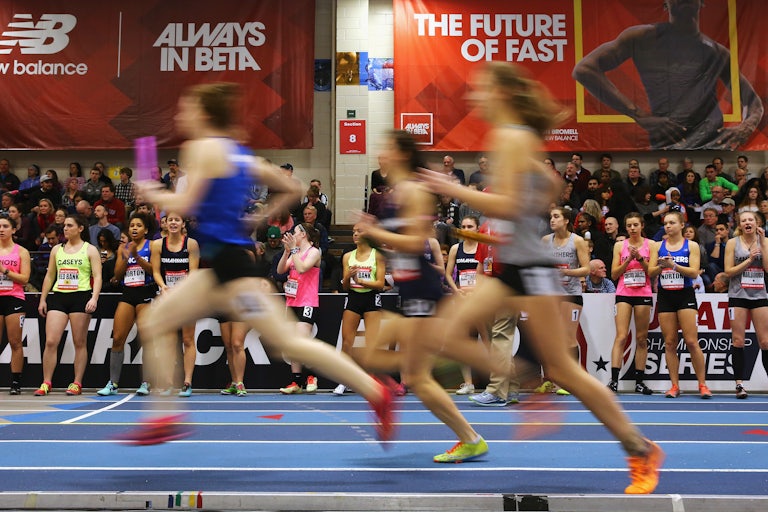 Three young women runners run by, blurred, on an indoor track, as other young runners and spectators look on.